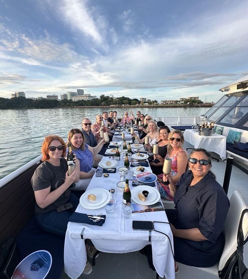 A long table on the deck of a boat, surrounded by people enjoying a meal together outdoors with the open water in the background