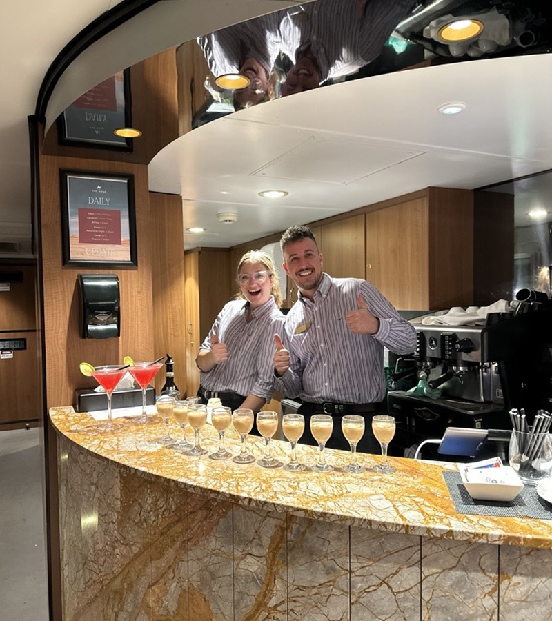Two smiling bartenders giving thumbs up behind a curved bar filled with many glasses of wine, inside the train lounge