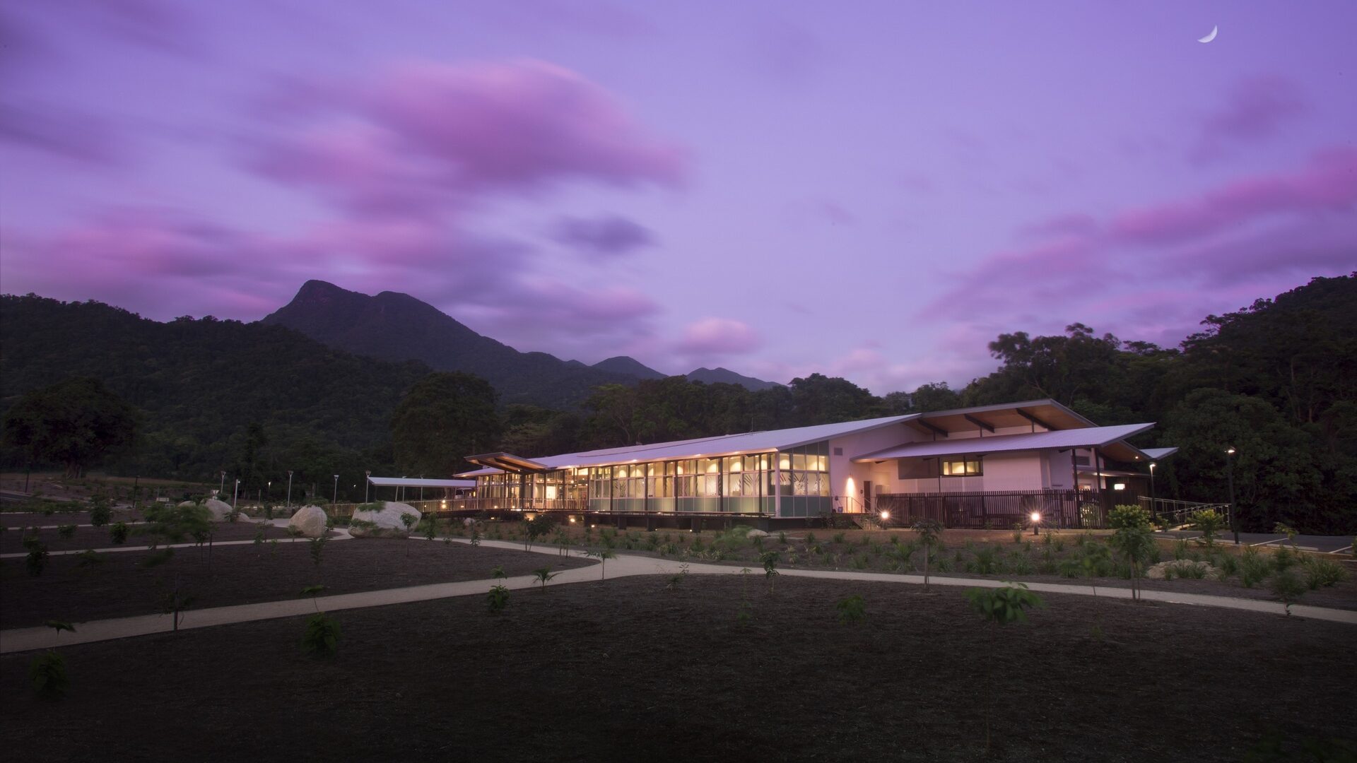 Mossman Gorge Cultural Centre Hero Shot - 1 A wide building cast in purple hues at sunset with a purple and pink sky above