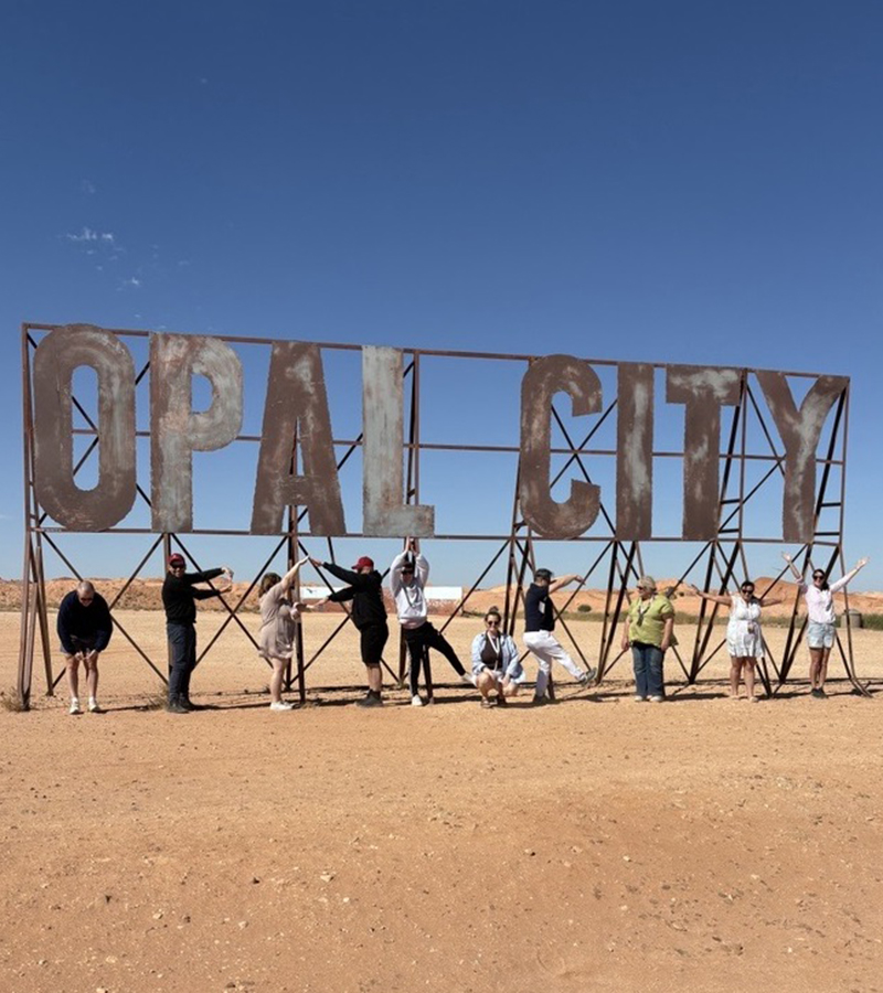Group of people standing on red outback soil in front of large sign made of metallic scaffolding that reads opal city