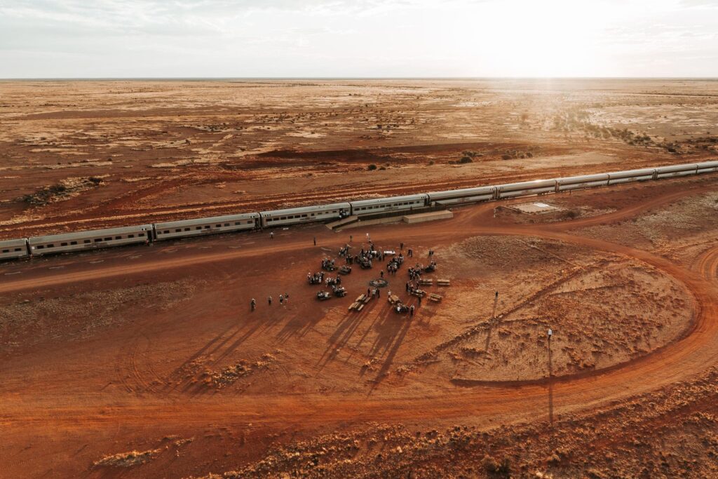 Overhead view of a long train stopped on wide red outback plains, with an elegant outdoor dinner setting arranged beside the carriages on the dirt