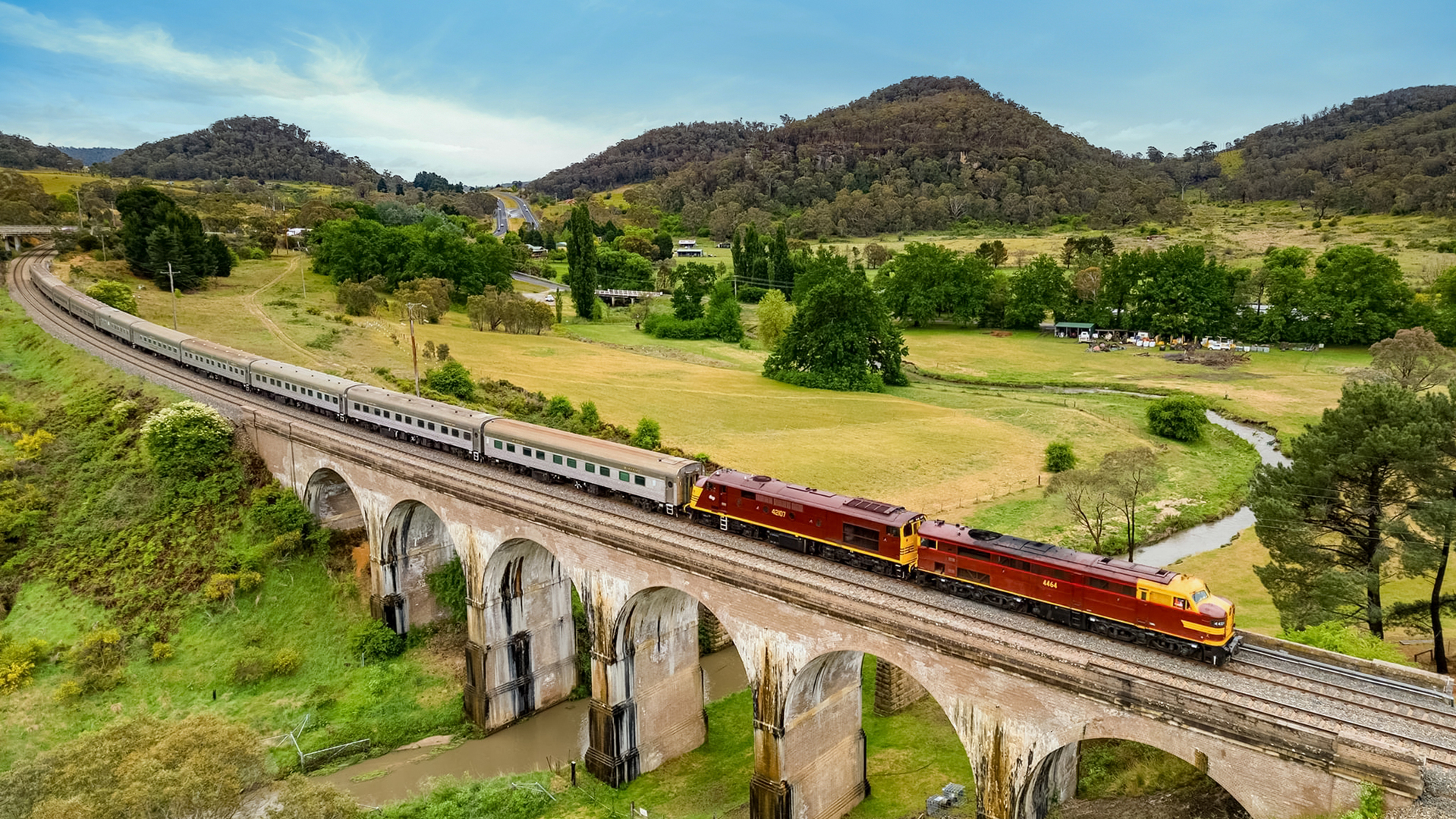 A train travels over a tall bridge with lush green hills in the background